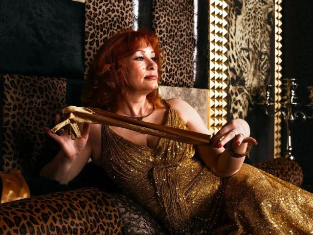 MomAimy-hot with red hair, wearing a gold, sparkly dress, holding a gold whip, sitting on a leopard-print couch, looking slightly annoyed. Boobs visible, sexy.
