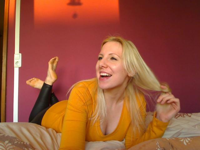 JuliaGold: morena, rubia, acostada en la cama, remera naranja de manga larga, leggings negros, sonriendo, sosteniendo el cabello, pies descalzos, pared rosa, luz naranja, juguetona, sexy, linda..