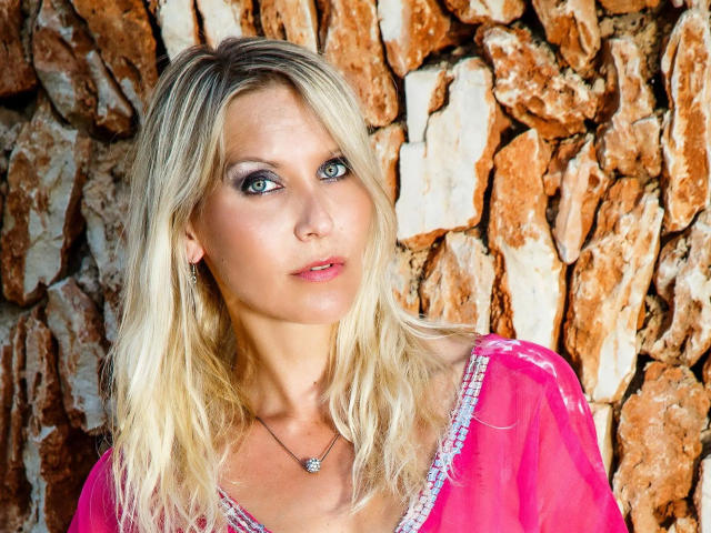 JuliaPink-hot heeft blond haar, blauwe ogen en draagt een roze, doorschijnende top met paarse randen, en staat voor een ruwe, rotsachtige muur. Haar borsten zijn klein en zichtbaar..