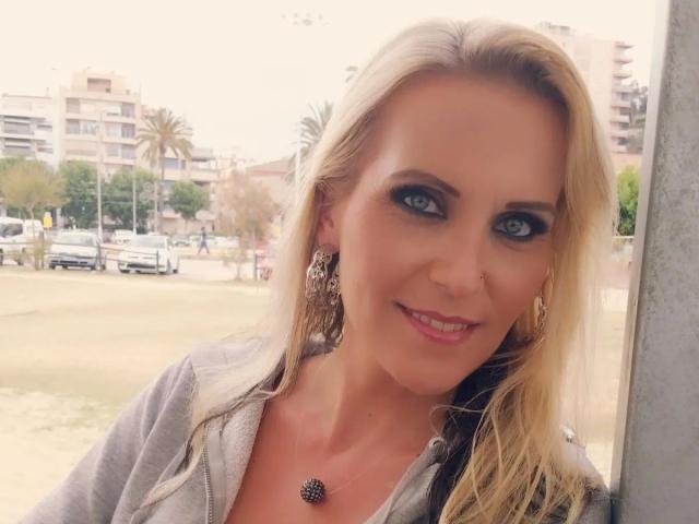 JuliaPink-hot a les cheveux blonds, les yeux bleus, et porte un sweat à capuche gris avec un collier noir orné d'épines. Elle sourit, laissant apparaître son rouge à lèvres rose, et porte des boucles d'oreilles travaillées..