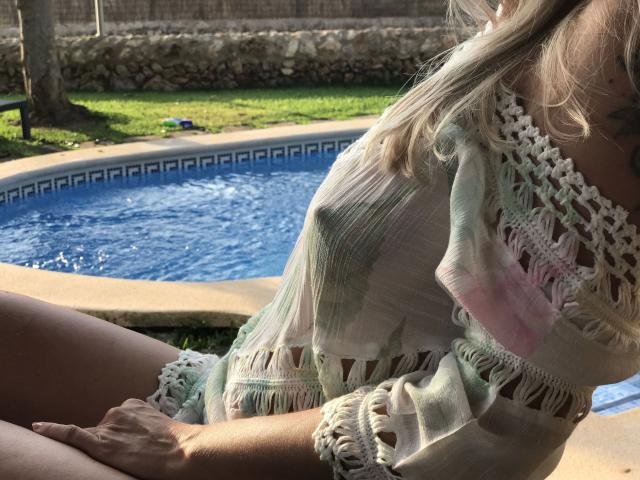 SexyClaudi-hot with her tits barely visible under a fringed, pastel-colored top, sitting by a blue pool, sunlight on her blonde hair, and her hand on her thigh.