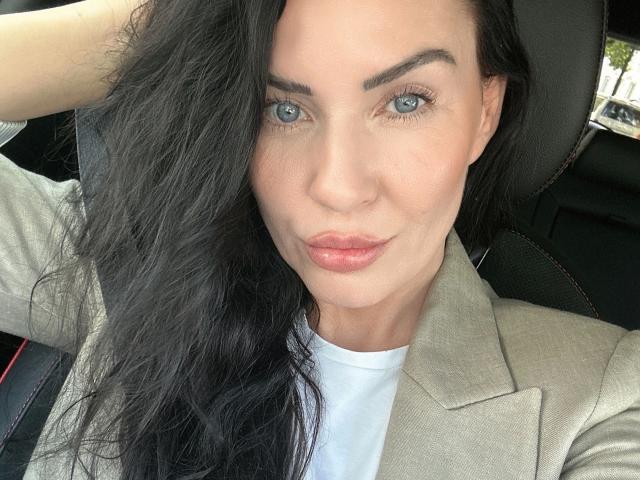 Malina-Lay, con occhi azzurri, capelli neri ondulati e labbra rosa, indossa un blazer beige, una camicia bianca e si siede su una sedia nera, guardando la fotocamera con una mano tra i capelli..