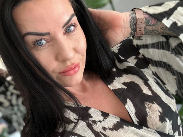 Malina-Lay, con capelli neri, occhi azzurri e labbra rosa, indossa una camicia a righe nere e bianche, mostrando un tatuaggio sul polso, con una mano tra i capelli. Il suo seno è parzialmente visibile..