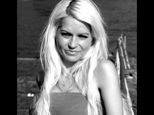 Black-and-white photo of jackylafey-hot with long, platinum blonde hair, small tits, and a cute, slightly annoyed smile. She's wearing a strapless top, and has a necklace.