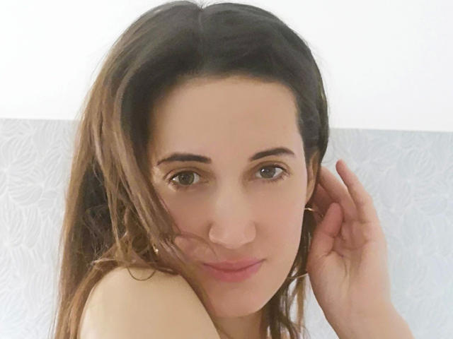 Lucilla69-hot with brown hair, green eyes, and pink lips, looking at camera, hand near ear, wearing small gold hoop earrings, light skin, soft smile, no visible clothes.