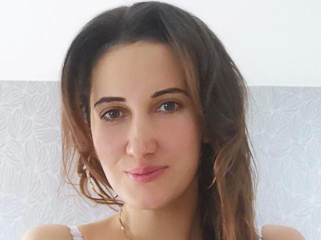 Lucilla69-hot has brown wavy hair, blue eyes, and pink lips, wearing a white tank top, with a slight smile. She's a cute, attractive woman with a fair skin tone.