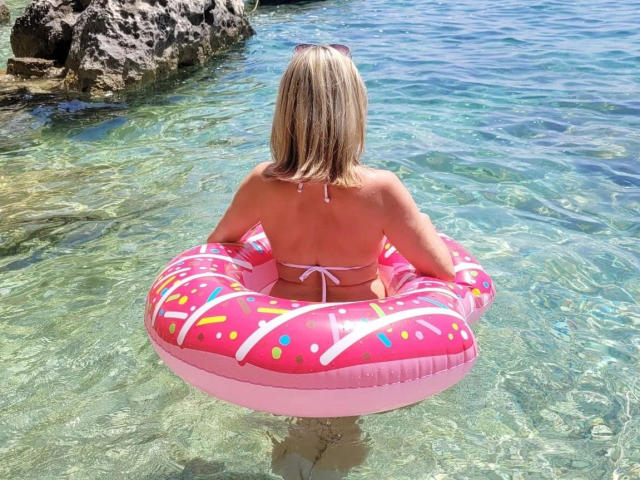 VIPLara-hot with blonde hair, back to camera, wearing a pink, donut-patterned inflatable, in clear, shallow water, pink bikini top visible, sunglasses on head, rocky shore in background.