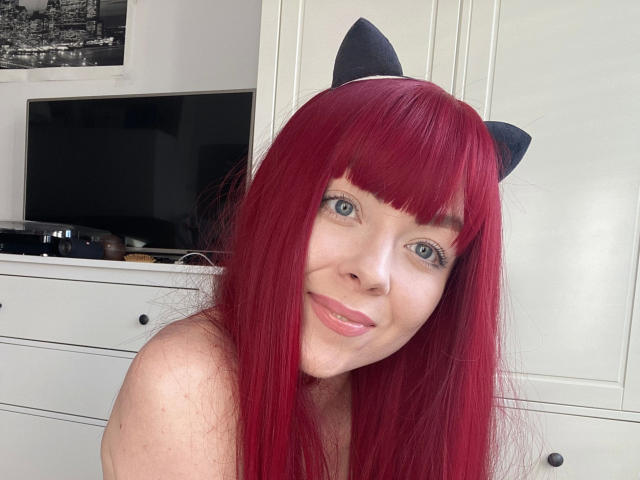 Jessie: Mit feuerrotem Haar, Katzenohren, blauen Augen und einem süßen Lächeln, wobei ihre nackten Schultern zu sehen sind, hat sie einen hellen Hautton und blickt in die Kamera..