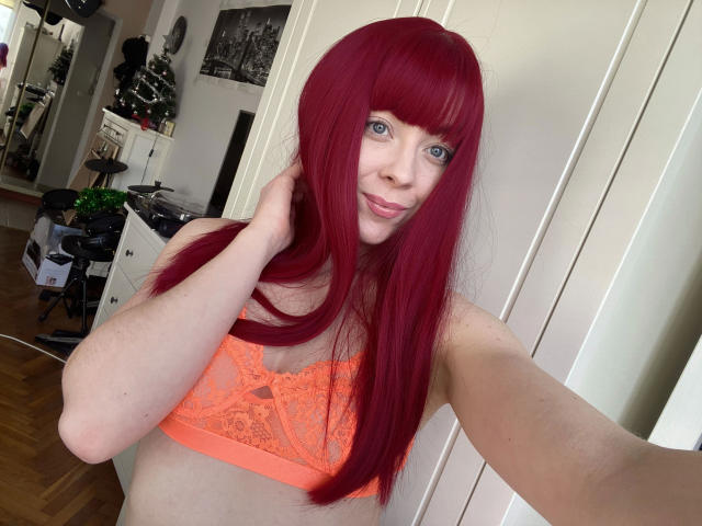 Jessie : Cheveux roux flamboyants, yeux verts, soutien-gorge en dentelle orange, souriant à la caméra, montrant ses seins, avec sa main touchant ses cheveux, dans une pièce bien éclairée..