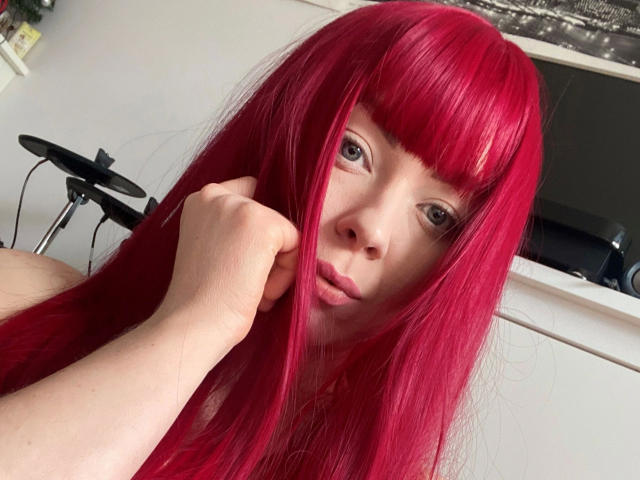 Jessie: Capelli rosso fuoco e luminosi, con una mano che tiene i capelli, guarda la fotocamera con un leggero sbuffo, pelle chiara e occhi verdi, in un ambiente interno informale..