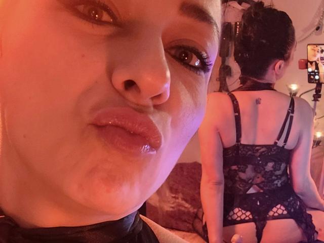 Mary-Licious with glossy lips, wearing black lace lingerie, showing her back with a tattoo on her upper right butt cheek, in a dimly lit room with warm red lighting.