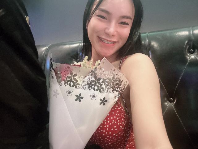 NaughtyAzumi69-hot with black hair, smiling, holding a bouquet of flowers with white and black floral patterns, wearing a red polka-dot dress, seated on a green leather couch.