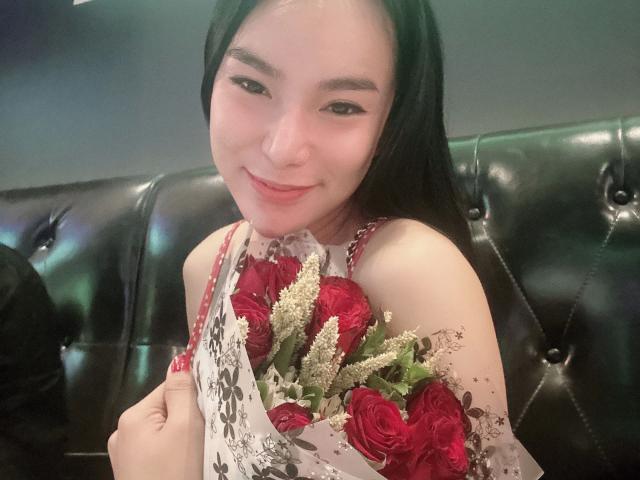 NaughtyAzumi69-hot with black hair, smiling, holding a bouquet of red roses and white flowers, wearing a floral dress, sitting on a black leather tufted seat.