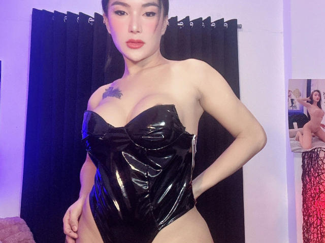 NaughtyAzumi69-hot with shiny black latex bodysuit, showing tits, tattoo on chest, hand on hip, standing, black curtain, mirror reflecting NaughtyAzumi69-hot in red lace, bedroom.