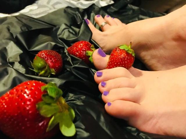 Os pés de Circea, com os dedos pintados de roxo, repousam sobre um tecido de cetim preto, com várias morangos vermelhas e folhas verdes espalhadas ao redor. Ela usa um anel no dedão direito..