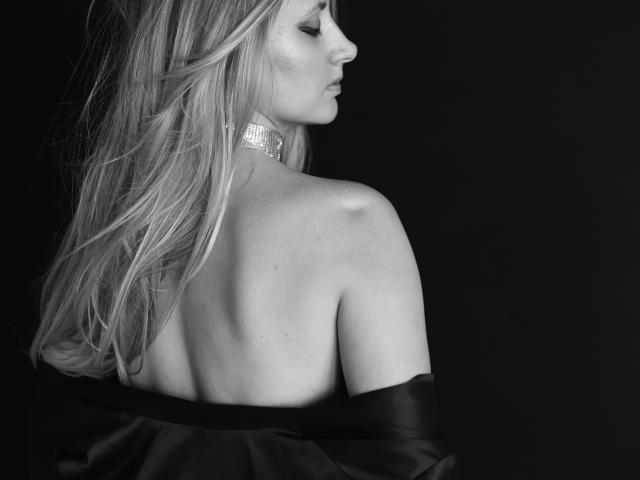 Circea, belle avec de longs cheveux blonds, dos nu, robe noire brillante à épaules dénudées, collier scintillant, vue de profil, fond noir, monochrome, élégante, sexy, peau lisse, expression douce..