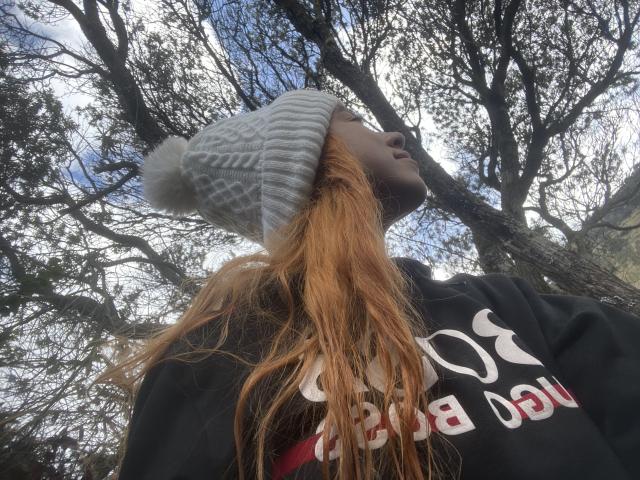 JuliaGarciax with long red hair, wearing a gray knit hat and black hoodie, looking up at tall, leafless trees under a blue sky. Her face is partially in shadow.