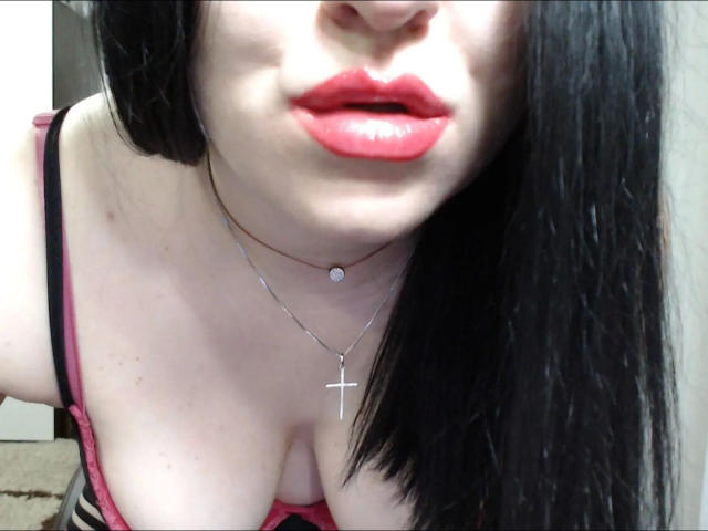 YourErica-hot with glossy red lips, black straight hair, silver cross necklace, pink and black bra, showing cleavage and slightly open mouth, leaning forward.