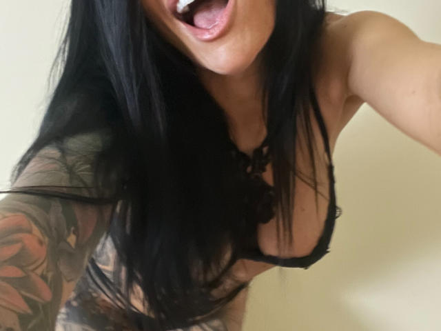 AndraD has long black hair, her mouth open with a white dildo in it, wearing a black lace bra, showing tattoos on her arm, and has large tits.