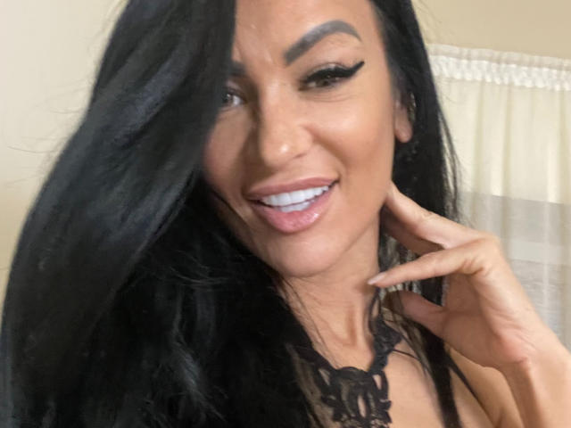 AndraD has black hair, thick eyebrows, and is smiling with white teeth. She's wearing black lace and has a hand near her face. Her makeup includes black eyeliner.