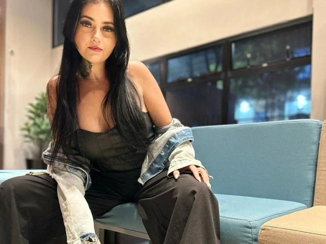 LaisFantasy-hot with long black hair, off-shoulder black top, black pants, and denim jacket, sitting on blue chair, looking at camera with slight smile. Her tits are small.