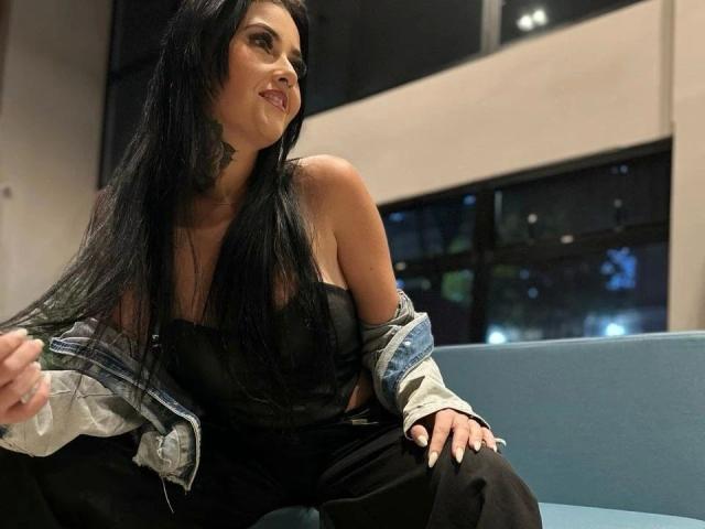 LaisFantasy-hot with long black hair, tan skin, and small tits, sitting on a blue couch, wearing a black strap top and denim jacket, smiling.
