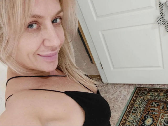 Alessandra93-hot with blonde hair, blue eyes, and pink lips, smiling at the camera, wearing a black spaghetti strap top, showing her bare shoulders and upper chest.