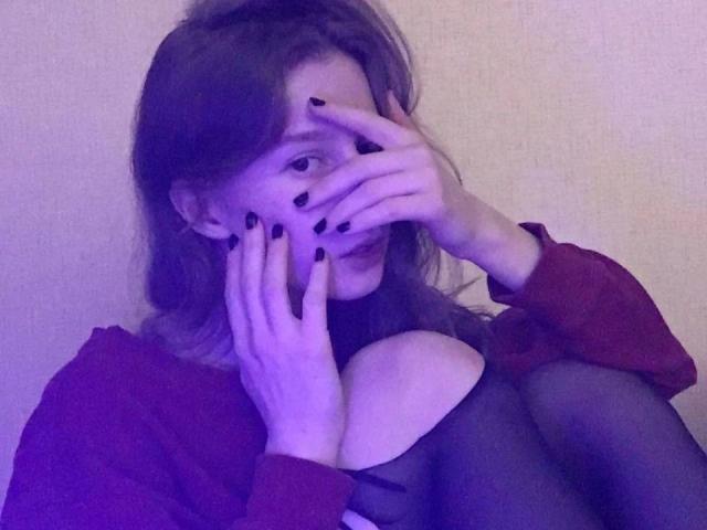 Enigma-hot with black nail polish covers her face, showing her boobs in a red shirt and black tights, sitting with knees up. Purple lighting.