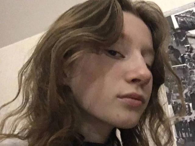 Enigma-hot has wavy brown hair, pale skin, and a serious expression with slightly parted lips. Her face is centered in the photo, with a black-and-white wall in the background.
