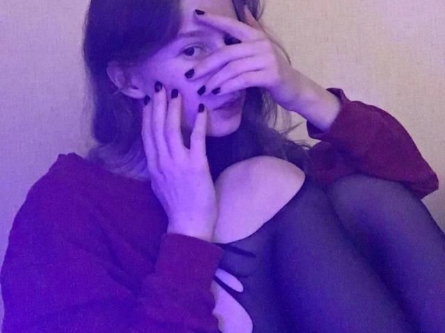 Enigma-hot with black nails, covering her mouth, wearing a red jacket, black cut-out dress, and sheer tights, sitting with knees up, in purple-lit, soft-focus image.