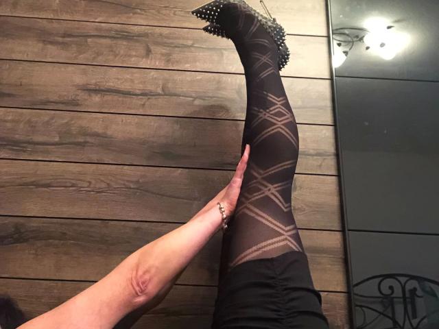 GeilesLuder-hot's leg is raised, showing black, patterned stockings with diamond shapes. A hand with a ring touches her leg. She's wearing black, studded heels.