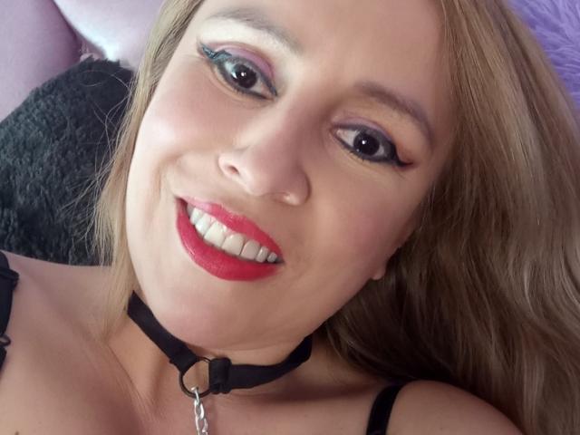 A MadissonS tem cabelo loiro, sombra azul, batom vermelho, colar preto com corrente, está sorrindo para a câmera, mostrando dentes brancos, vestindo uma blusa preta com alças, e tem pele clara..