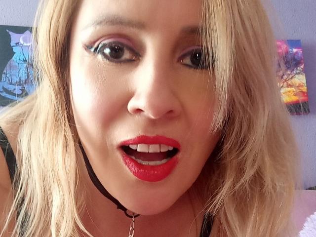MadissonS tiene el cabello rubio, lápiz labial rojo y delineador de ojos negro, con un collar y una cadena negros, y está mirando a la cámara con la boca ligeramente abierta..