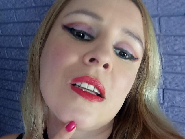 MadissonS a un fard à paupières rose, un eye-liner noir, un rouge à lèvres rouge et un vernis à ongles rose, avec des cheveux blonds et une bouche légèrement ouverte, touchant son menton..