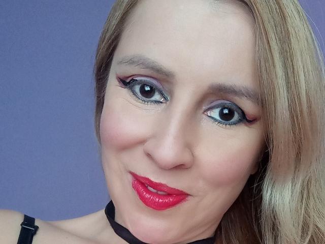 MadissonS tiene ojos azules grandes, lápiz labial rojo, delineador de ojos negro y cabello rubio. Lleva un collar negro, está sonriendo con una cara linda. Sus senos no son visibles..