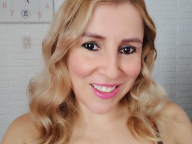 A MadissonS tem cabelo loiro cacheado, delineador preto, batom rosa e um sorriso adorável. Seus seios não estão visíveis. Ela tem um tom de pele claro. Ela está olhando para a câmera..
