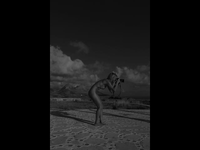 Lulu-4-you is naked, bending forward with a camera in hand, showing her tits and ass, standing on a stone surface under a dark, cloudy sky. Black and white.