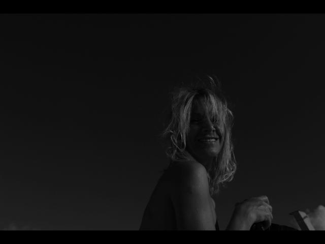 Lulu-4-you has messy blonde hair, a dark, shadowy face, and is smiling while holding something with her right hand, in a black-and-white, low-light photo.