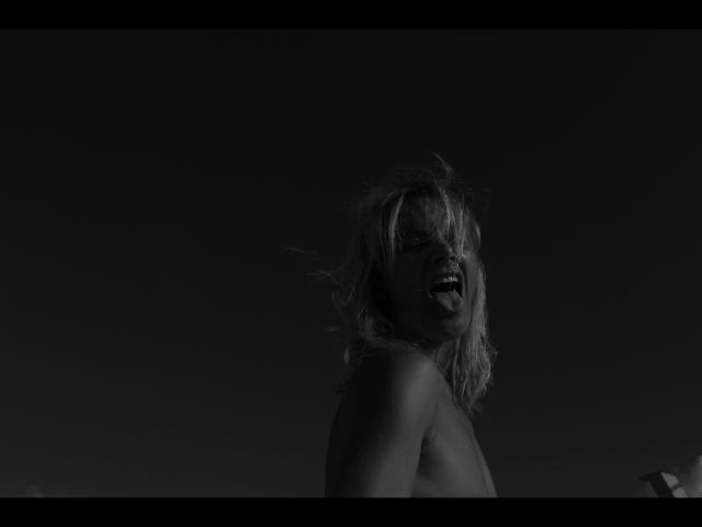 Black-and-white photo of Lulu-4-you with messy blonde hair, tongue out, topless, showing small tits, dark background, and soft light highlighting her playful expression.
