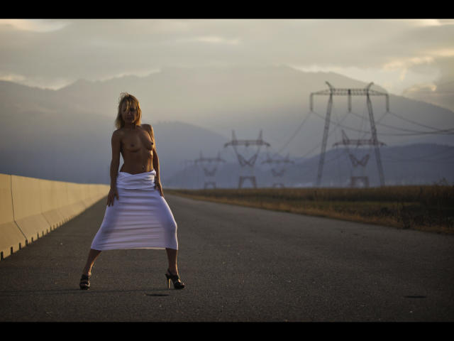 Lulu-4-you, topless with tits, wearing a white skirt, standing on a road, hands on hips, in black heels, with power lines and mountains in the background.