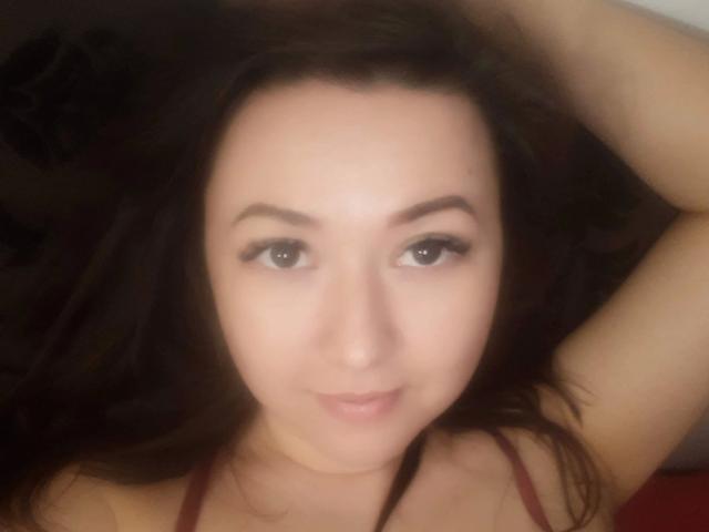 A selfie of Goldmoon-hot with dark brown hair, light skin, and brown eyes, lying down with a slight smile, showing her upper chest and arm. Boobs visible.