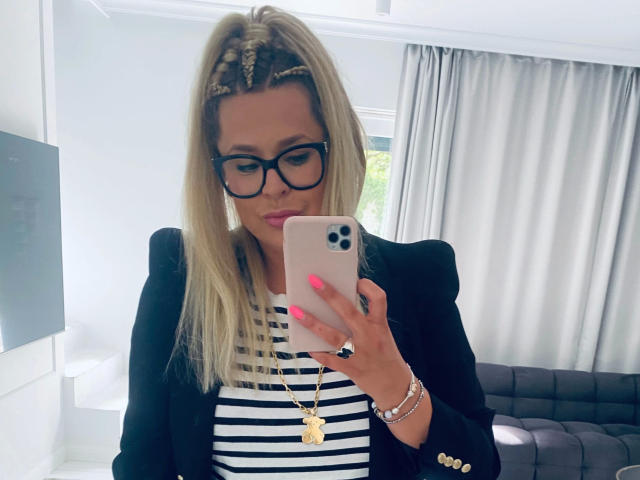 Diana-extasy with blonde hair, black glasses, striped shirt, black blazer, pink nails, gold necklace, holding iPhone, smiling, white-walled room, gray curtains, black sofa.