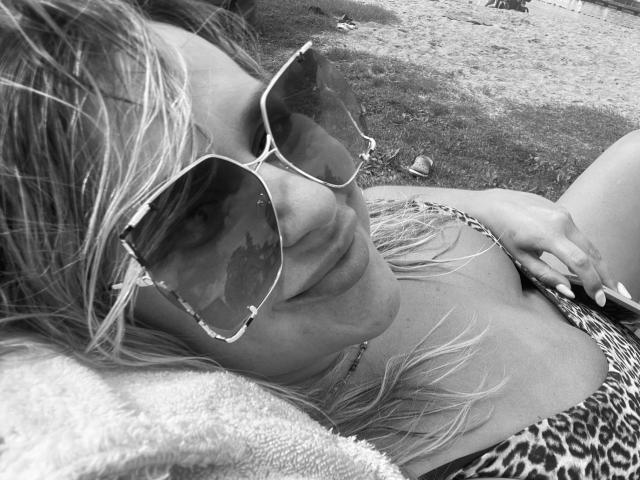 Diana-extasy lying on a towel, wearing cat-eye sunglasses and a leopard print bikini, with blonde hair and a necklace, looking relaxed. Her hand is on her chest.