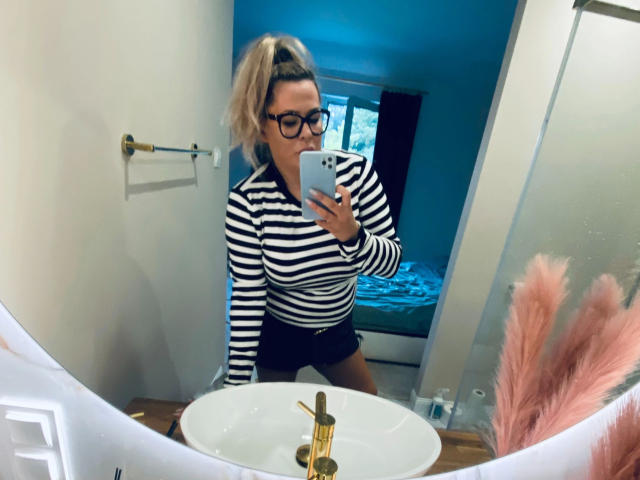Diana-extasy with blonde ponytail, black glasses, striped shirt, and black shorts, taking a mirror selfie in a bathroom. Her small tits are visible under the shirt.
