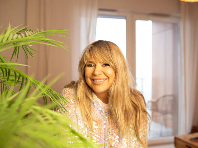 Diana-extasy with blonde hair, smiling, wearing a sparkly top, standing in front of a green plant, sunlight on her face, soft focus, warm indoor lighting.