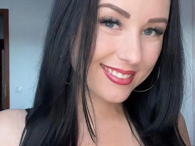 MilaJane-hot has black hair, blue eyes, red lipstick, and is smiling with hoop earrings. She's wearing a black top, showing her tits and ass isn't visible.