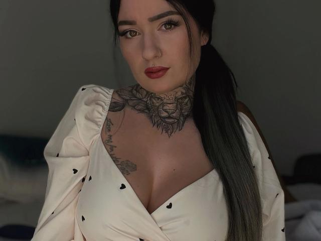 Claudiliciousxx-hot with dark tattoos around her neck and chest, wearing a white dress with heart patterns, has red lipstick, and long black hair in a ponytail. Boobs visible.