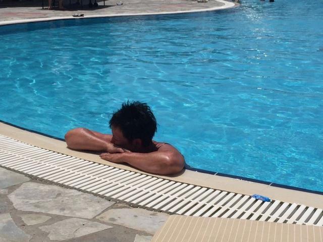 Sexy-Milf with black hair, shirtless, arms folded, resting by blue pool, tan tiled edge, stone pattern floor, small blue toy on tile.