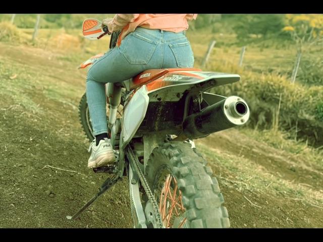 BlackAngeel's ass is up on a dirt bike, wearing tight blue jeans and white sneakers, with a pipe on the bike's back. The bike's orange and gray.