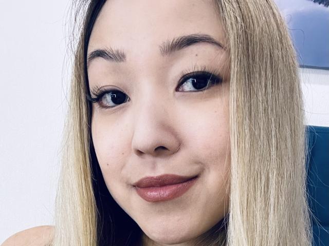 RenyLime's face, blonde hair, thick eyebrows, black eyeliner, long lashes, subtle smile, pinkish-brown lipstick, light skin, slight freckles, looking to the side, soft light.